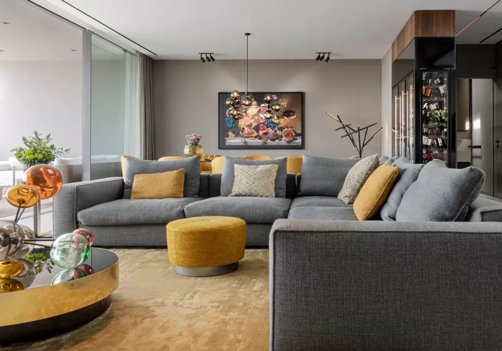Modernes Wohnzimmer mit großer grauer Eckcouch, goldgelben Akzenten und einer skulpturalen Designerlampe. Ein runder Couchtisch mit farbigen Glaskugeln und eine elegante Weinvitrine ergänzen das stilvolle Interieur.