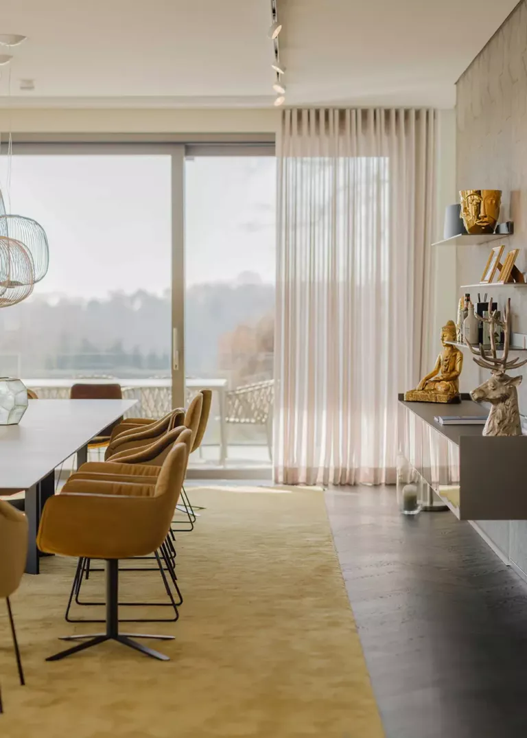 Elegantes Esszimmer mit langem Tisch, senffarbenen Stühlen und großen Fenstern mit Blick auf die Terrasse. An der Wand befinden sich dekorative Regale mit goldenen Skulpturen.