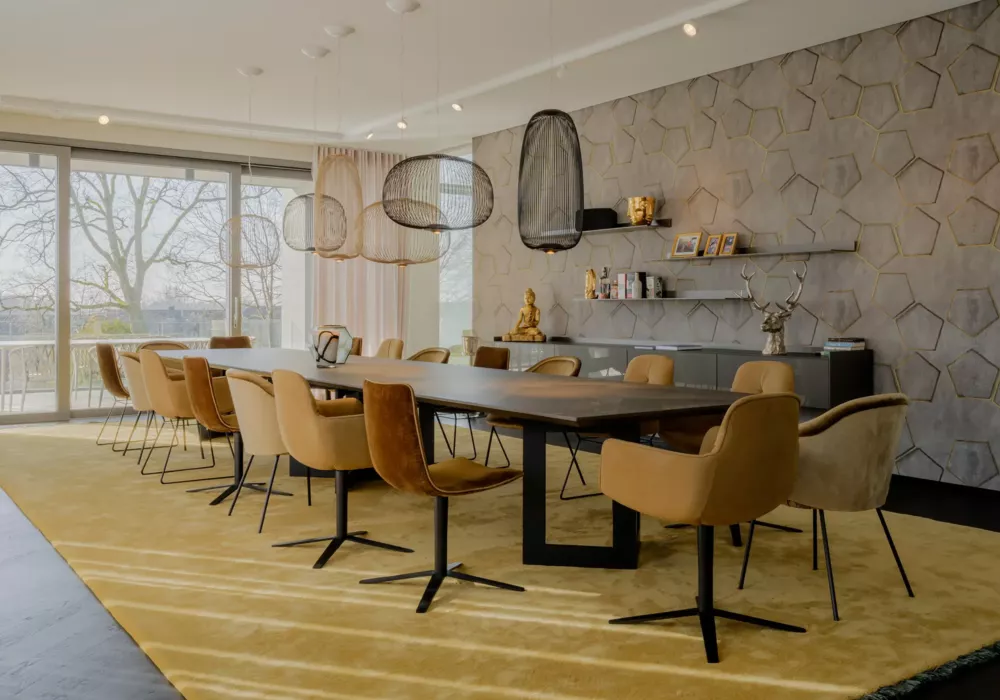 Modernes Esszimmer mit großem Holztisch, senffarbenen Stühlen und geometrischer Wandverkleidung. Hängeleuchten und bodentiefe Fenster mit Blick auf die Terrasse sorgen für eine helle Atmosphäre.