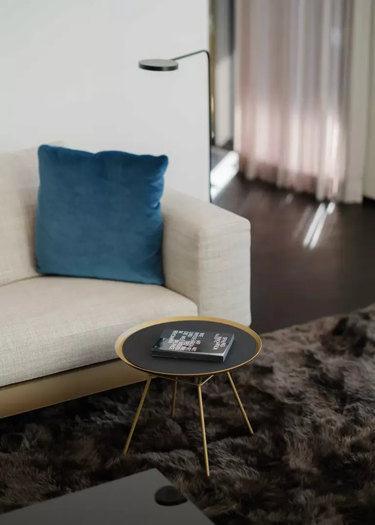 Elegante Wohnzimmerecke mit cremefarbenem Sofa, blauem Kissen und einem schwarzen Beistelltisch mit goldenen Beinen. Ein Buch liegt auf dem Tisch, daneben eine moderne Stehlampe und ein flauschiger Teppich.