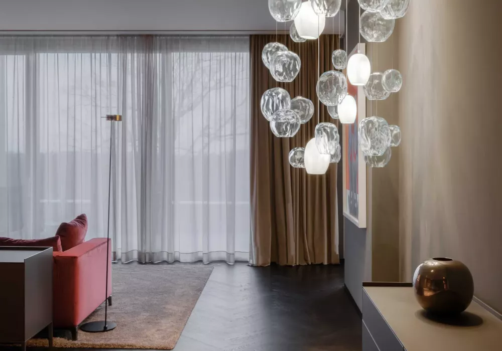 Modernes Wohnzimmer mit einem roten Sofa, einer schlanken Stehlampe und einem bodentiefen Fenster mit transparenten Vorhängen. Eine auffällige Glas-Pendelleuchte hängt an der Decke, während eine bronzefarbene Vase auf einer Konsole steht.