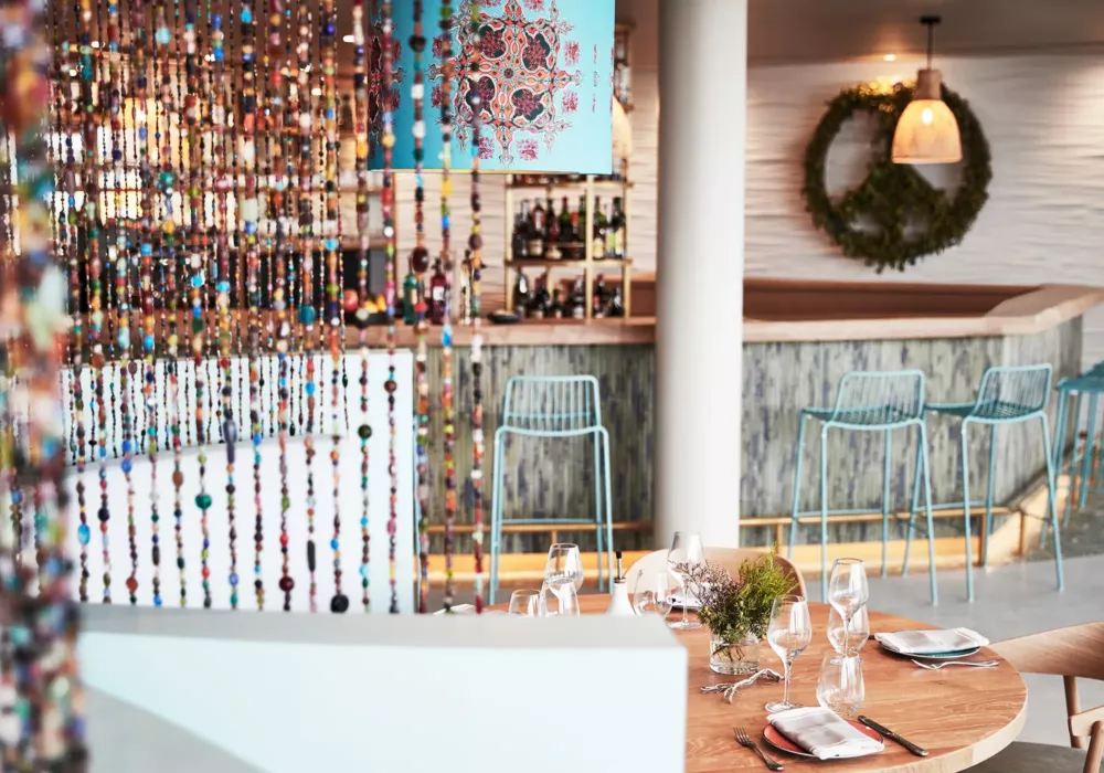 Stilvolles Restaurant im 7Pines Resort Ibiza mit hölzernem Esstisch, buntem Perlenvorhang und einer modernen Bar im Hintergrund