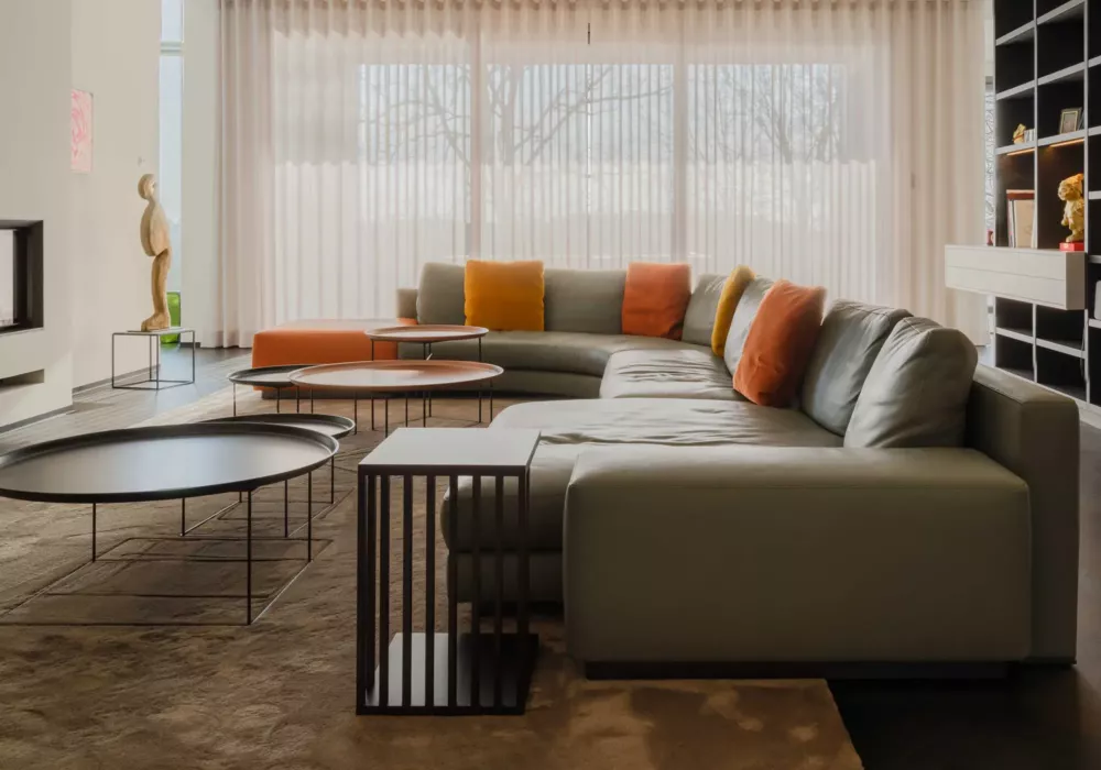 Modernes Wohnzimmer mit einer großen grauen Eckcouch, farbigen Kissen, mehreren runden Couchtischen und bodentiefen Fenstern mit Vorhängen.