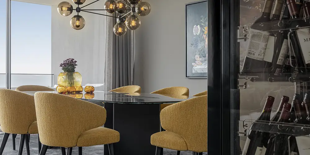 Modernes Esszimmer mit schwarzem Glastisch, senfgelben Polsterstühlen, stilvollem Kronleuchter und Weinschrank im Vordergrund.