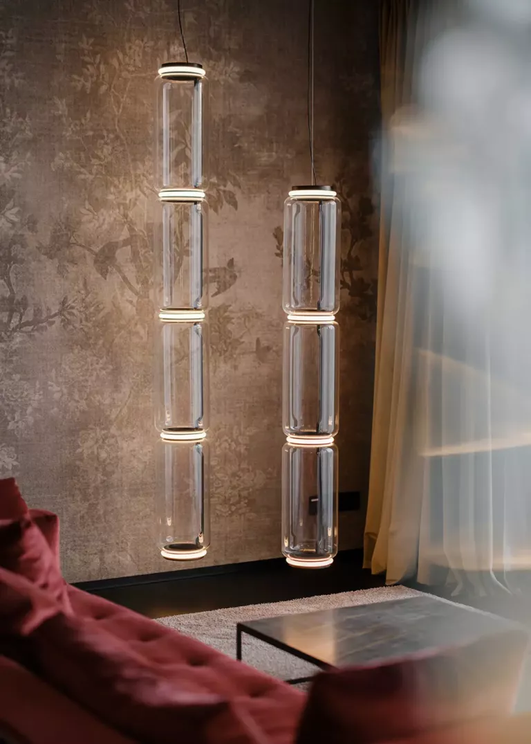 Elegante Pendelleuchten aus Glas mit zylindrischem Design, hängend vor einer gemusterten Tapete, neben einem roten Sofa und einem Metall-Couchtisch.