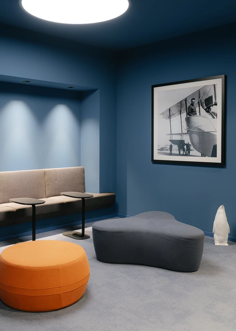 Moderner Lounge-Bereich mit tiefblauen Wänden, gepolsterter Sitzbank und stilvollen Hockern in Orange und Grau. Ein Schwarz-Weiß-Fotodruck eines historischen Flugzeugs ergänzt das Design.